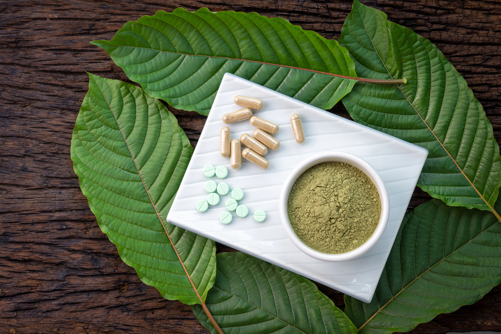 Compliant kratom product pages Kratom leaves, powder in a bowl, gel capsules, and small green tablets arranged on a white ceramic tray