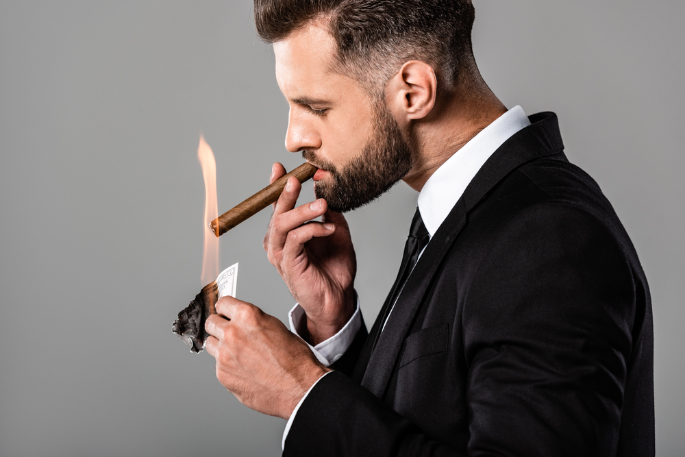 Tobacco shops selling cigars Man in black suit lighting a cigar with a burning banknote against a gray background
